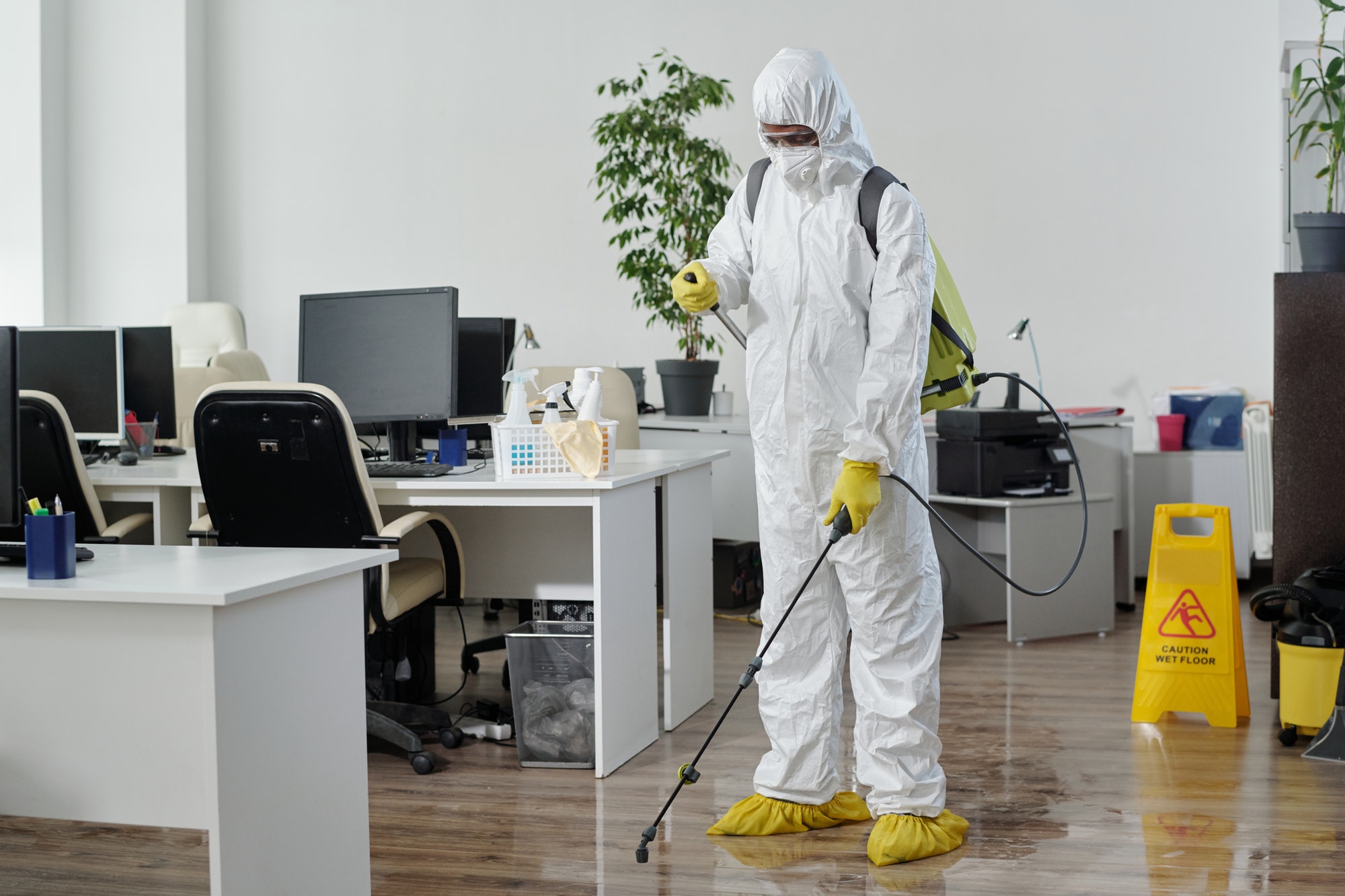 Male worker of cleaning service in hazmat suit disinfecting floor in office